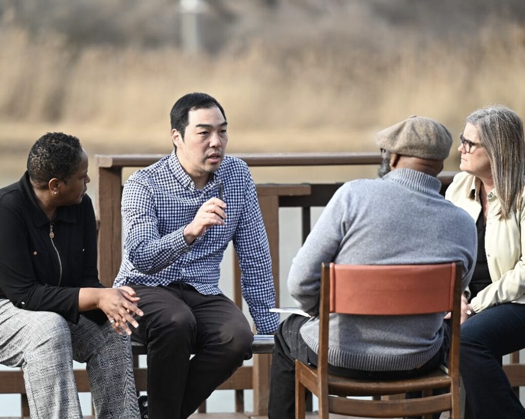 Four adults sit outdoors in a circle, engaged in a serious conversation on a wooden deck.
